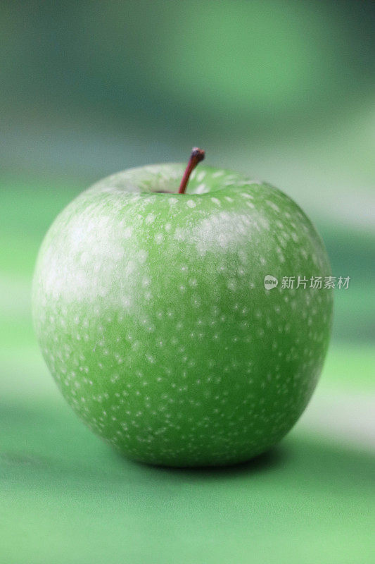 个体的特写图像，绿色的史密斯苹果(Malus domestica)，有茎，光滑的，有斑点的果皮，斑驳的绿色背景，重点在前景